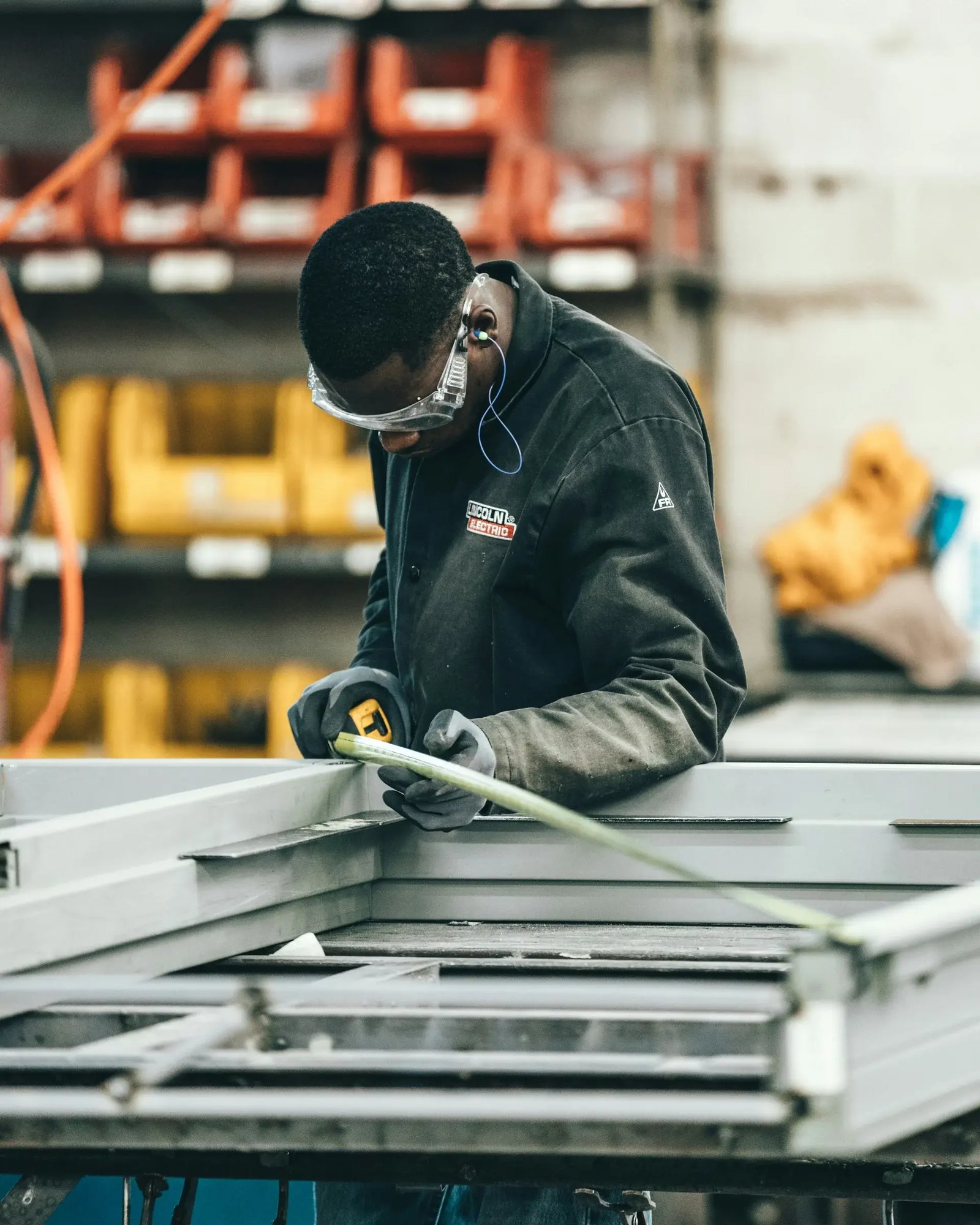 Metal fabricator in safety glasses and gloves measuring a steel workpiece with a tape measure in a fabrication shop.