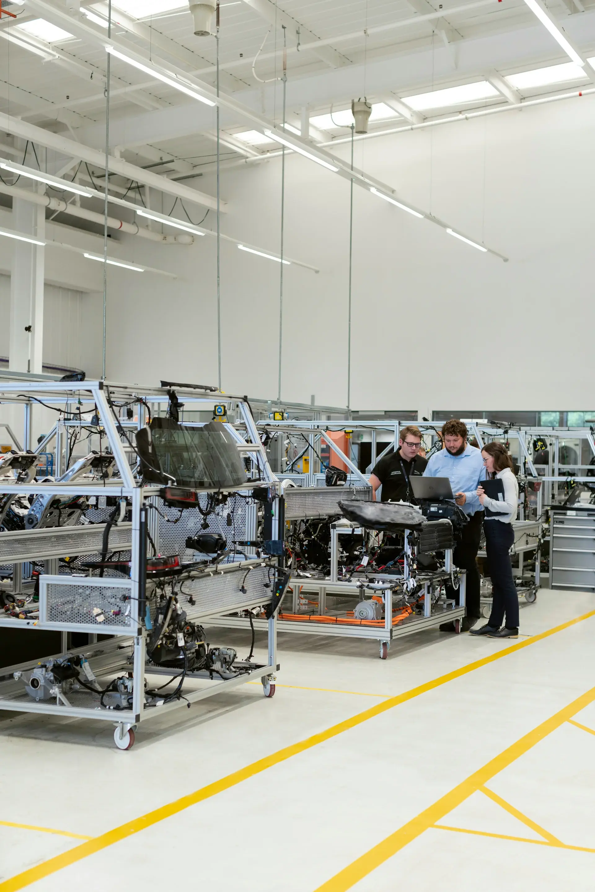 Three engineers reviewing data on a laptop beside vehicle assembly stations in a clean automotive engineering facility.